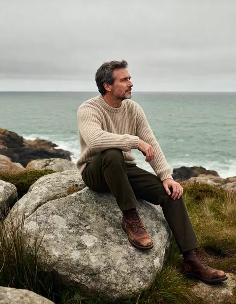 Homme en pull en maille assis sur un rocher face à la mer